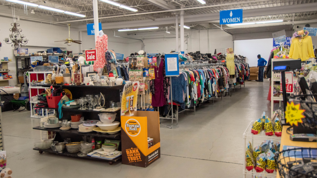 The interior of a large thrift store showing long racks of colorful clothing, shelves stocked with dishes, glassware, and household items, and blue department signs including a "Women" section marker. A "Clearance Sale" sign and a Tootsie Roll donation bank are visible among the displays.