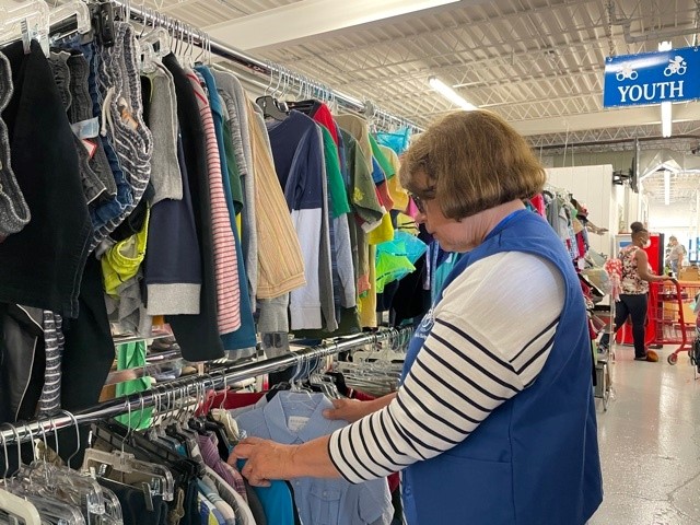 A volunteer in a blue vest and striped shirt sorts through clothing on a rack inside what appears to be a thrift store or donation center, with a "Youth" section sign visible in the background.