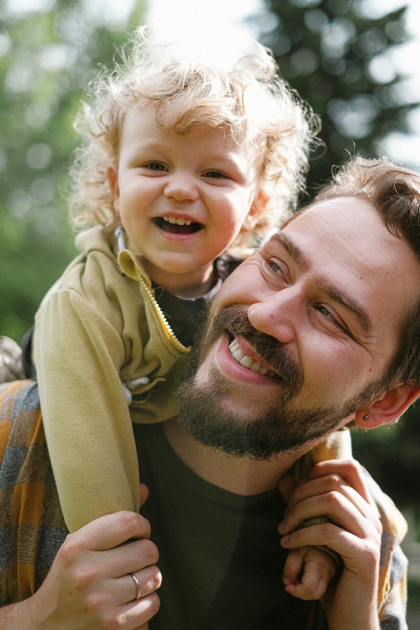 A bearded man smiles warmly while a laughing toddler with curly blond hair sits on his shoulders outdoors against a blurred green background.