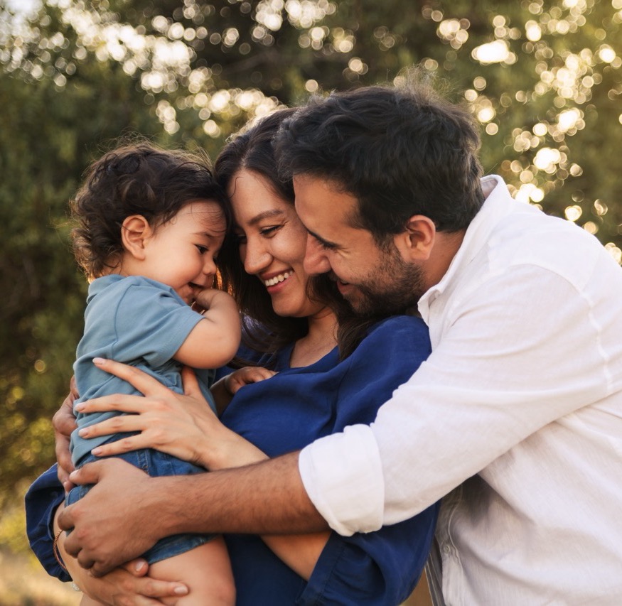 A joyful family moment with a father, mother in a blue dress, and their one-year-old son in blue clothes, hugging and smiling in a grassy natural setting with a tree in the background
