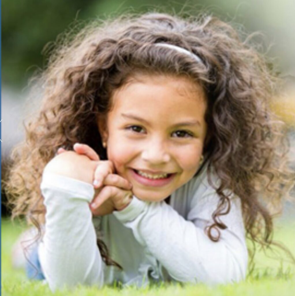 A smiling young girl with curly hair lying on green grass, representing the children and families helped by the Society of St. Vincent de Paul Northeast Ohio.