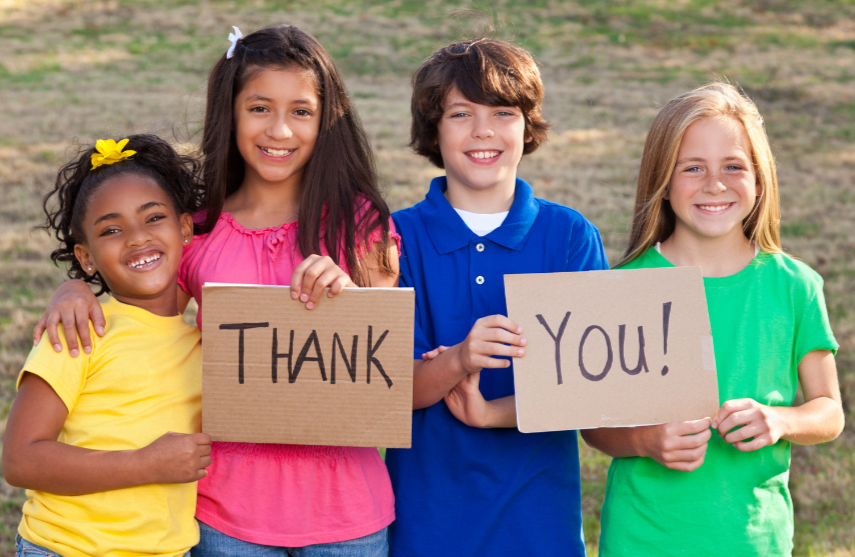Kids wearing colorful shirts, smiling outside holding sign